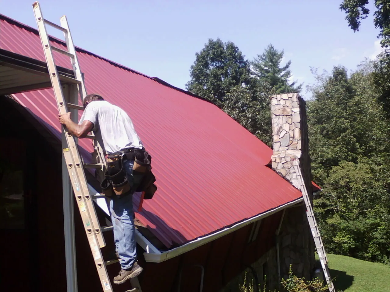 Roofer on a ladder beside a red metal roof and stone chimney — Soffit & Fascia Repair by Roofing Brothers in Haverhill
